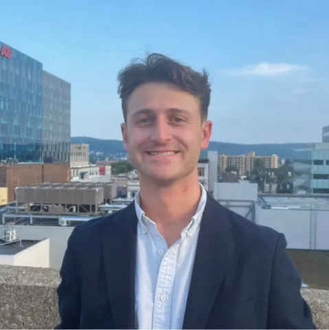 Trevor Tormann wearing a white button-up shirt and black jacket smiling on top of a building in Allentown, PA.
