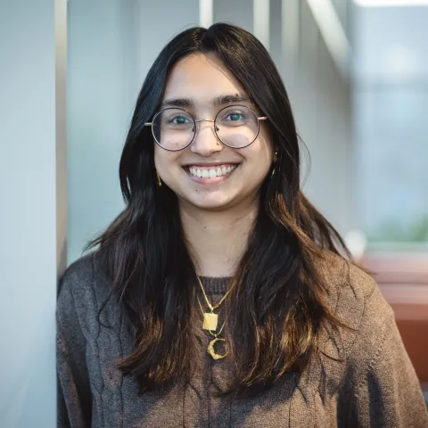 Shinam Hussain standing for her headshot wearing glasses and a brown sweater.